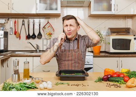 Perturbed Stress Young Man In Apron Sitting At Table With Vegetables, Talking On Mobile Phone, Cooki