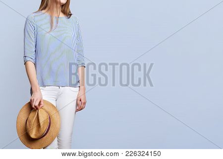 Cropped Shot Of Unrecognizable Female Wers Striped Blouse And White Trousers, Holds Straw Hat, Wears