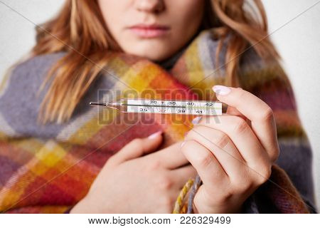 Cropped Shot Of Young Female Wrapped In Warm Coverlet, Holds Termometer Which Shows High Temperature