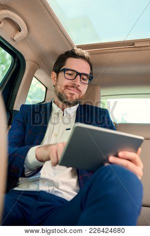 Smiling Young Businessman Working Online With A Digital Tablet While Sitting In The Backseat Of A Ca