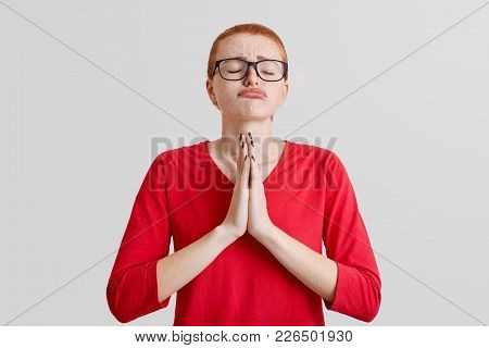 Horizontal Shot Of Hopeful Red Haired Woman With Shut Eyes, Presses Palms Together, Has Pleading Exp