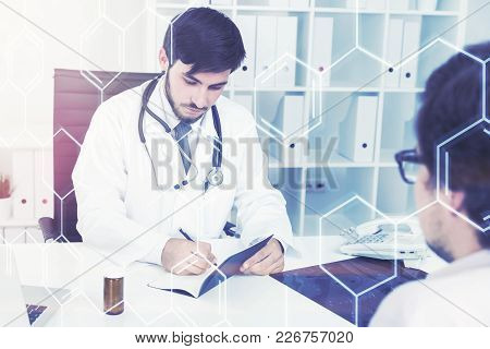 Bearded Young Doctor Is Talking To His Black Haired Patient In Glasses And Taking Notes. An Atomic G