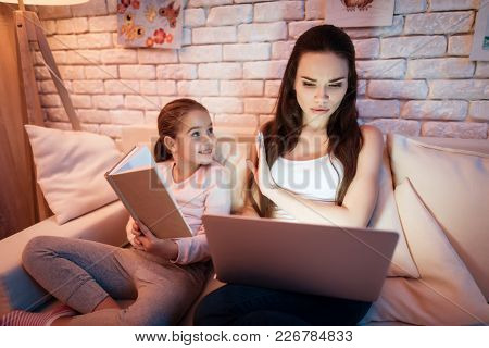 Little Daughter Wants Nother To Read Book But Mother Is Busy On Laptop At Night At Home.