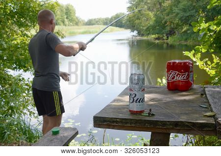 Sumy, Ukraine - August 10, 2019: Budweiser Bud Beer Cans Pack On Old Table And Fisherman At River On