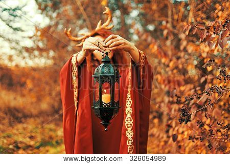 Woman In Long Red Dress With Deer Horns In Fall Forest. The Demon Of Fairy Tales In A Red Cloak With