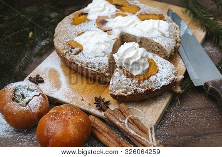 Winter Cake With Persimmon On A Wooden Board, Knife, Cinnamon Sticks, Star Anise, Spruce Branches