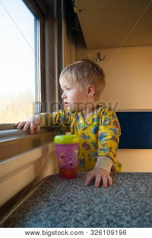 A Bright, Cute Three-year-old Boy Riding A Train Looks Out The Window, Behind Which A Dull Landscape