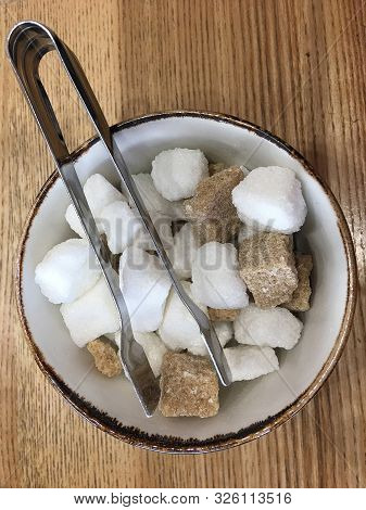 Top View Of A Bowl With Pieces Of White And Brown Sugar Cubes And With Sugar-tongs On A Wooden Table