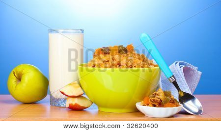 leckere Cornflakes in grüner Schale, Äpfel und Glas Milch auf Holztisch auf blauem Hintergrund