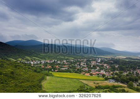 Small Village In A Valley. Hills, Mountains And Fields Around. Dark Cloudy Summer Day. Picture Taken