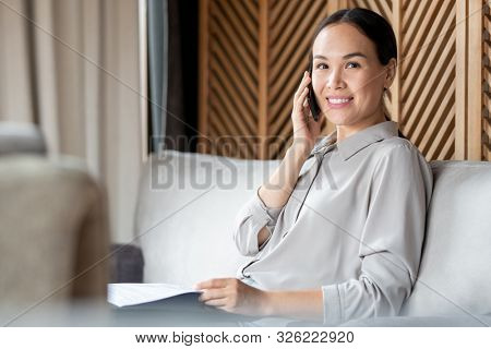Young successful female emloyee with smartphone and papers sitting on couch in cafe and consulting client