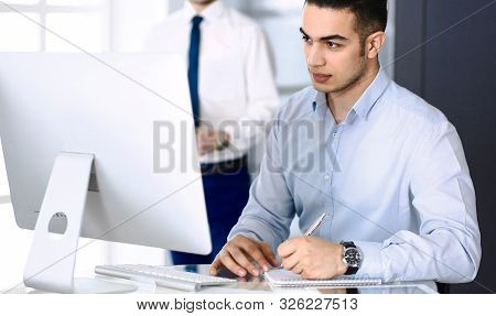 Businessman Working With Computer In Modern Office, Male Colleague At Background . Headshot Of Arab 
