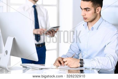 Businessman Working With Computer In Modern Office, Male Colleague At Background . Headshot Of Arab 
