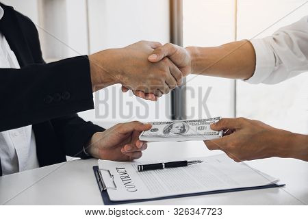 Two Corporate Businessmen Shaking Hands While One Man Places Money On Document In Office Room With C