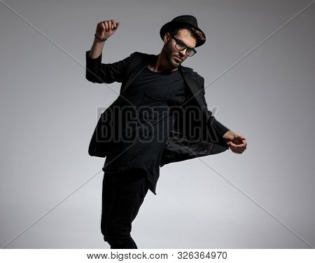 sexy casual man wearing hat and glasses standing and pulling his jacket aside with elegance against gray studio background