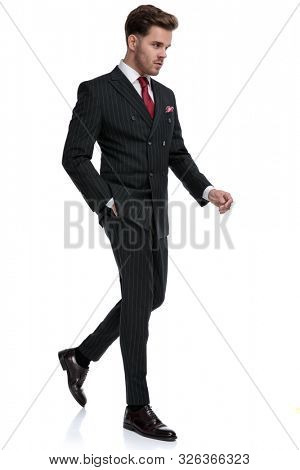 side view of elegant young businessman wearing double breasted suit and red tie, walking and looking to side, isolated on white background in studio