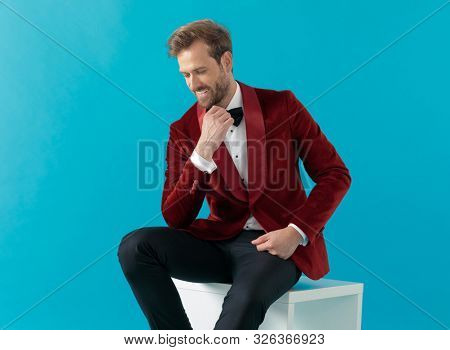 smiling fashion model wearing red velvet tuxedo, holding hands to chin, looking down and sitting on blue background