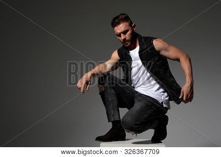 Thoughtful young man adjusting his jeans vest and squatting on a chair while wearing black jeans on gray studio background