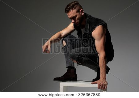 Handsome man squatting and getting off a chair while wearing a black jeans vest on gray studio background