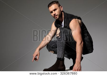 Tough man pointing down and squatting while wearing a black jeans vest and jeans on gray studio background