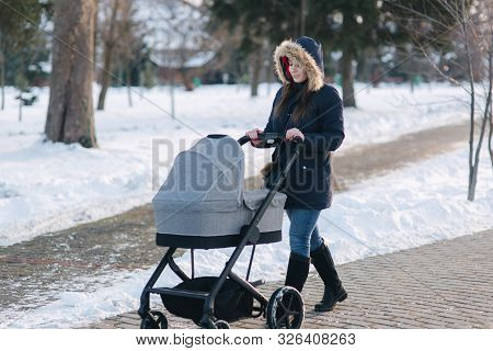Beautiful Mother Walking In The Park With Her Little Baby In Stoller. Woman Dressed In Blue Jaket Wi