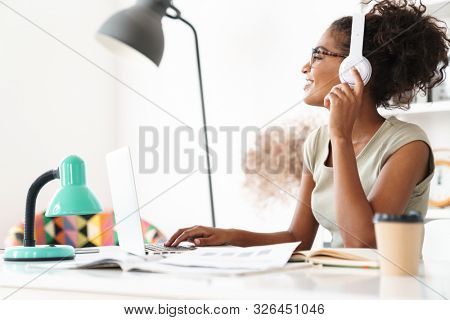 Image of a pretty happy positive optimistic young african woman sit in office using laptop computer indoors listening music with headphones.