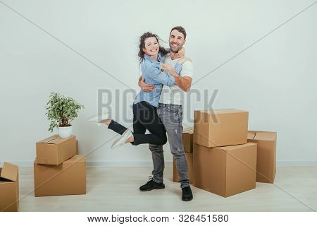 Couple Moving Into Their New House. Woman Is Very Glad And Is Hanging On Her Husbands Neck, Hugging 