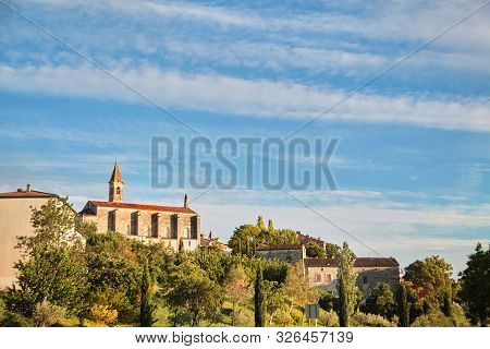 View Of  The Old Town Of Barjac, Southern France. Green Trees, Buildings And Stone Church With A Bel