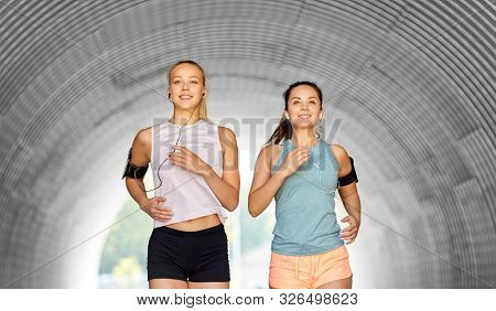 fitness, sport and healthy lifestyle concept - happy young women or female friends with earphones wearing armbands with smartphones and running outdoors
