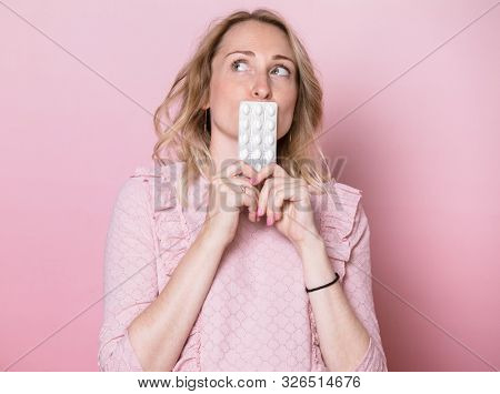 Young pregnant woman wearing pink dress holding blister pack of medicine tablets considering to take pills during pregnancy or not. Studio shot on pink background. Prenatal medication concept
