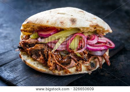 Traditional barbecue pulled beef burger offered with Carolina BBQ sauce, gherkin and onion rings as closeup on a burnt wooden board 