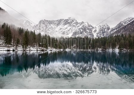 Lake With Green Forest And Snowy Mountains In The Background And Skyfall Effect. Italian Lake During