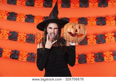 Image of sassy witch girl in black halloween costume showing horn fingers and holding carved pumpkin isolated over orange wall