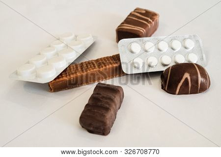 Chocolate Candies And Pills Lie Together On A White Table. Medicaments And Sweets.