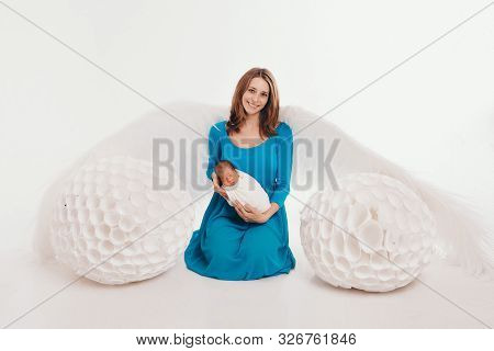 A Girl In A Blue Dress With Angel Wings Holding A Newborn Baby On A White Background. An Angel-woman