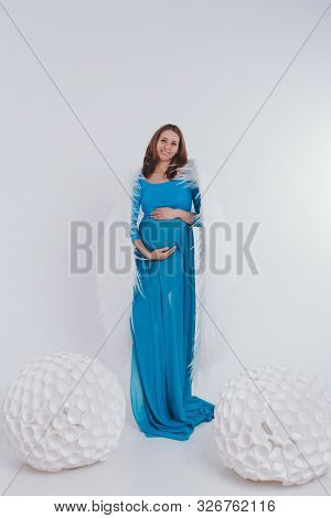 A Pregnant Girl In A Blue Dress With Angel Wings On A White Background. An Angel-woman Have Big Whit