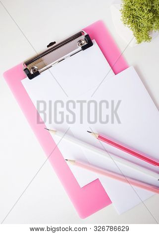 Three Pencils Lie On A Pink Clipboard And A Sheet Of Paper. In The Frame You Can See A Plant In A Ca