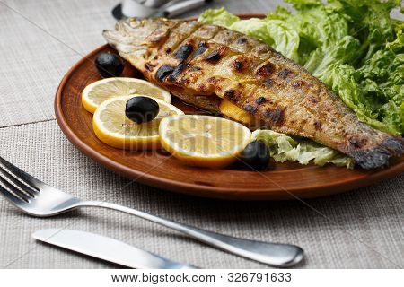 Grilled Trout In Brown Clay Bowl Close-up. Front View Of Plate With Grilled Trout Garnishe With Sala