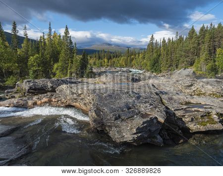 Beautiful Northern Landscape With Wild Glacier River Kamajokk, Boulders And Spruce Tree Forest In Kv