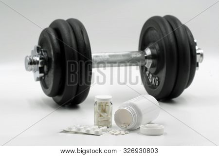 White Pills In Front Of Black Dumbbell Isolated On White Background. Concept Of Using Supplements Fo