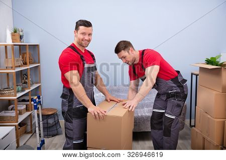 Portrait Of Two Smiling Young Male Movers In Uniform Packing Cardboard Boxes In Living Room
