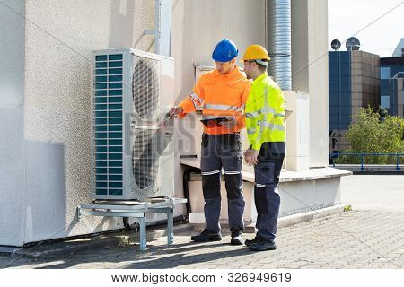 Two Electricians Men Wearing Safety Jackets Checking Air Conditioning Unit On Building Rooftop