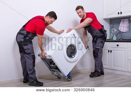 Two Young Male Professional Movers In Uniform Placing The Modern Washing Machine In The Kitchen