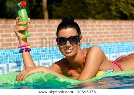 Bella donna sorridente posa sul materassino in piscina, bere cocktail.