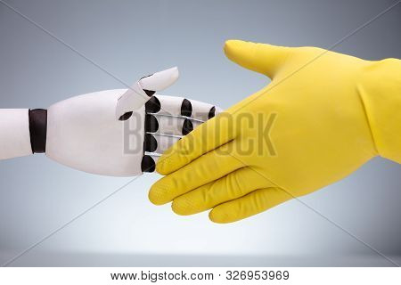 Close-up Of Robot And Janitor Wearing Yellow Glove Shaking Hands Against Gray Background