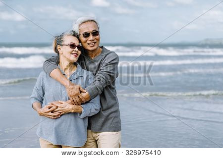 Asian Senior Couple Or Elderly People Walking And Siting At The Beach On Their Weekend Vacation Holi
