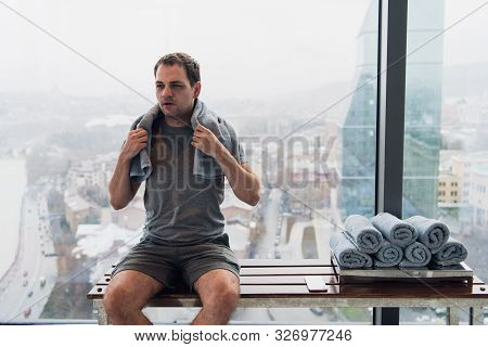 Young, Tired Man Sitting On Bench In A Gym. Holding Mobile Phone And Towel On His Head. Gym At Skysc