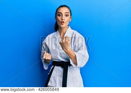 Beautiful brunette young woman wearing karate fighter uniform with black belt doing attack pose making fish face with mouth and squinting eyes, crazy and comical. 