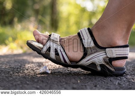 Woman Foot In Sandals Steps On A Shabby Wireless Headphones That Lies On A Asphalt Sidewalk, Outdoor