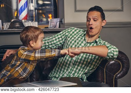 Young Father And His Stylish Little Son In The Barbershop In The Waiting Room.